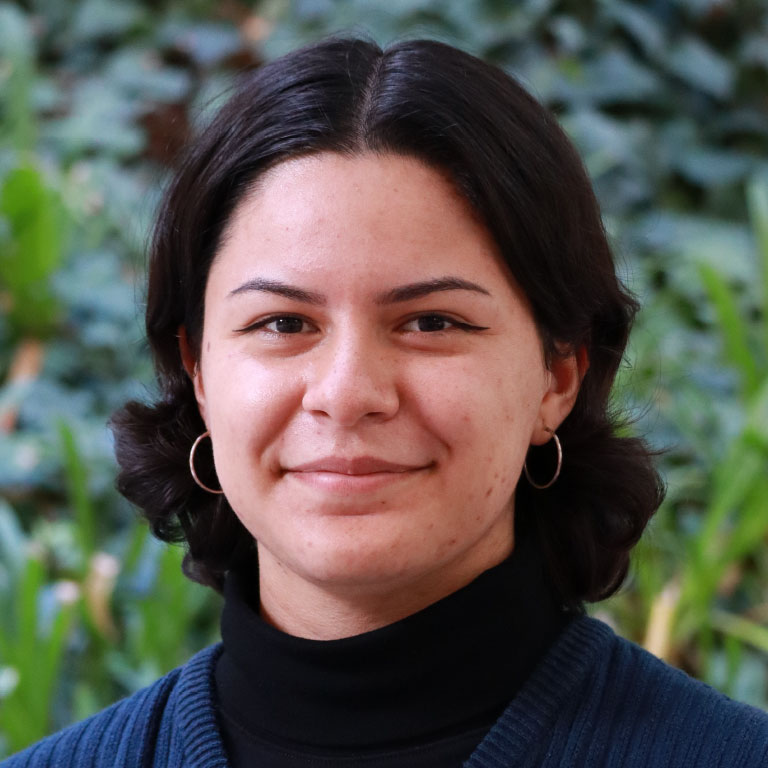 Headshot of scholar. Green plants behind scholar. Scholar has black short hair, gold earrings and is wearing a black turtle neck and blue sweater vest.