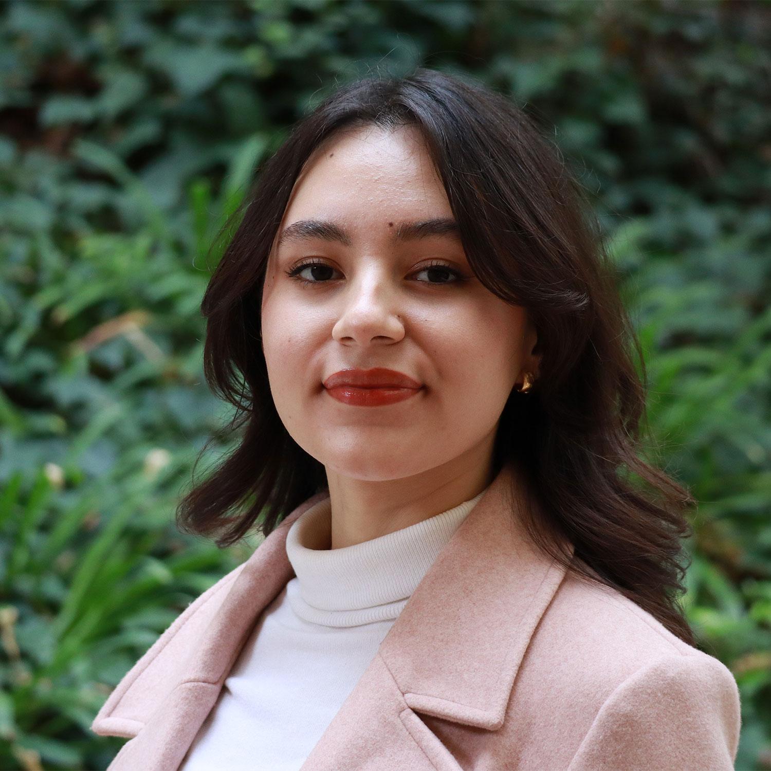 Headshot of scholar. Green plants behind scholar. Scholar has black short hair, gold earrings and is wearing a white turtle neck and pink blazer