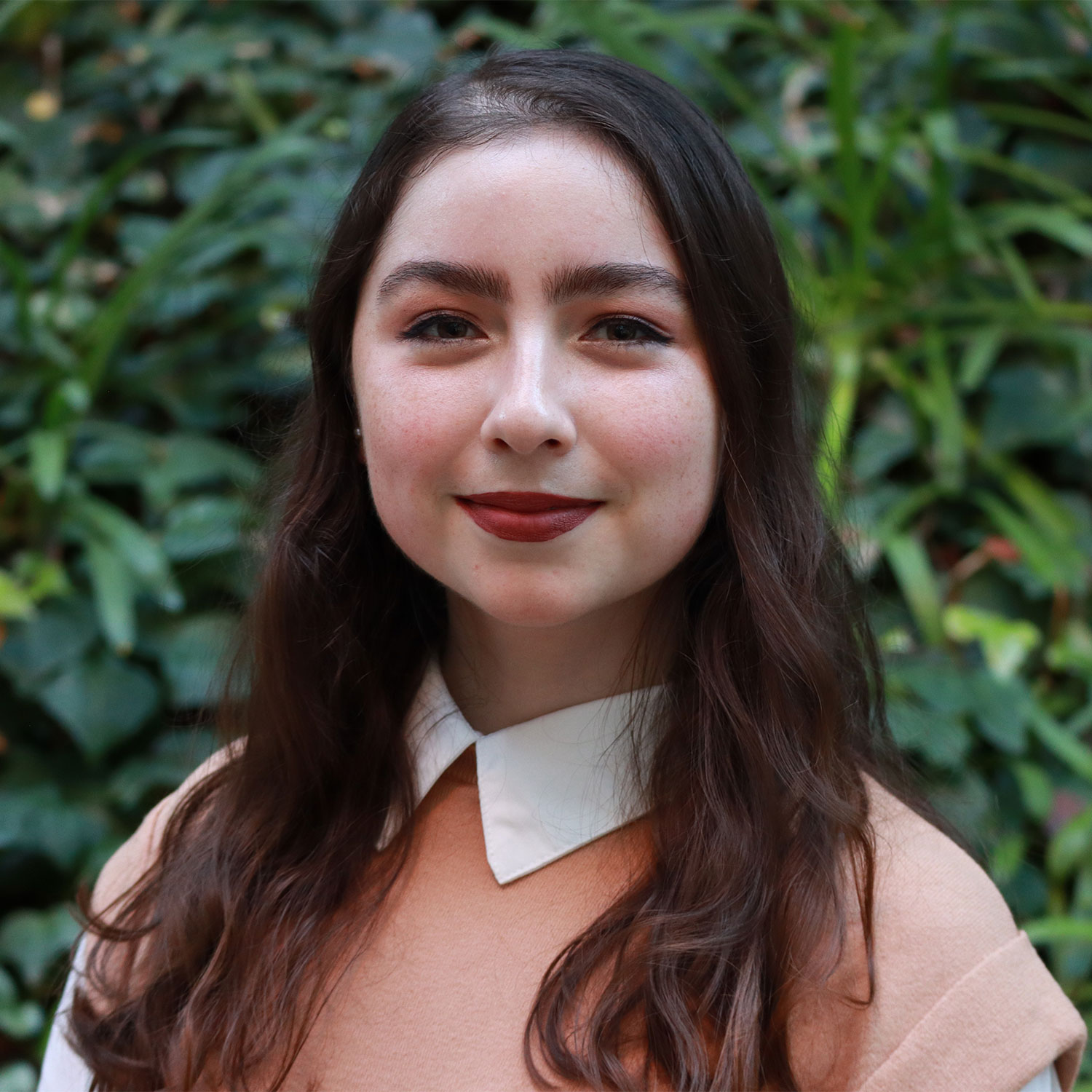 Headshot of scholar. Green plants behind scholar. Scholar has long brown hair, red lipstick. They are wearing a white collar shirt with a pink sweater on top.