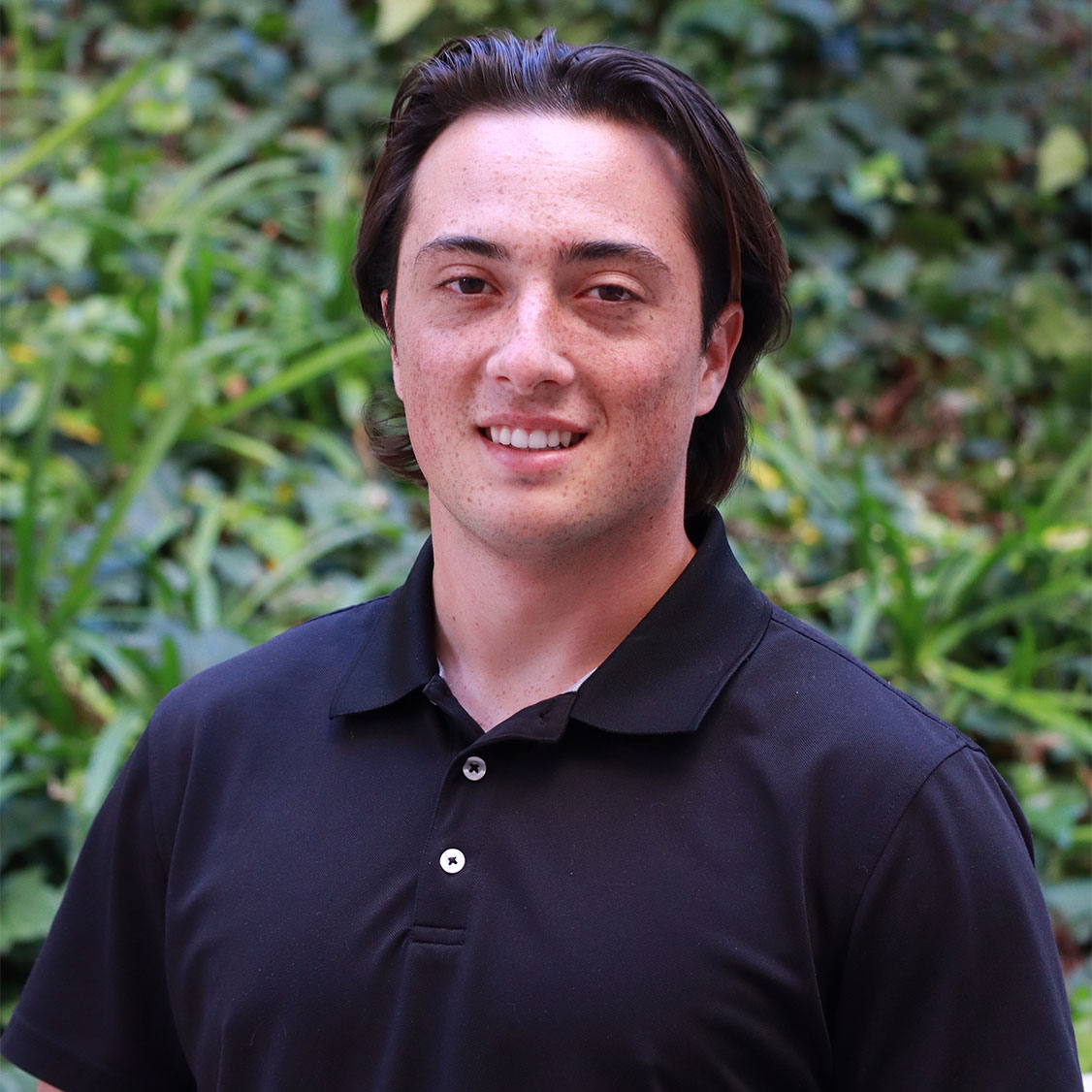 Headshot of scholar. Green plants behind scholar. Scholar has medium length brown hair. Freckles. Wearing a black button up polo shirt