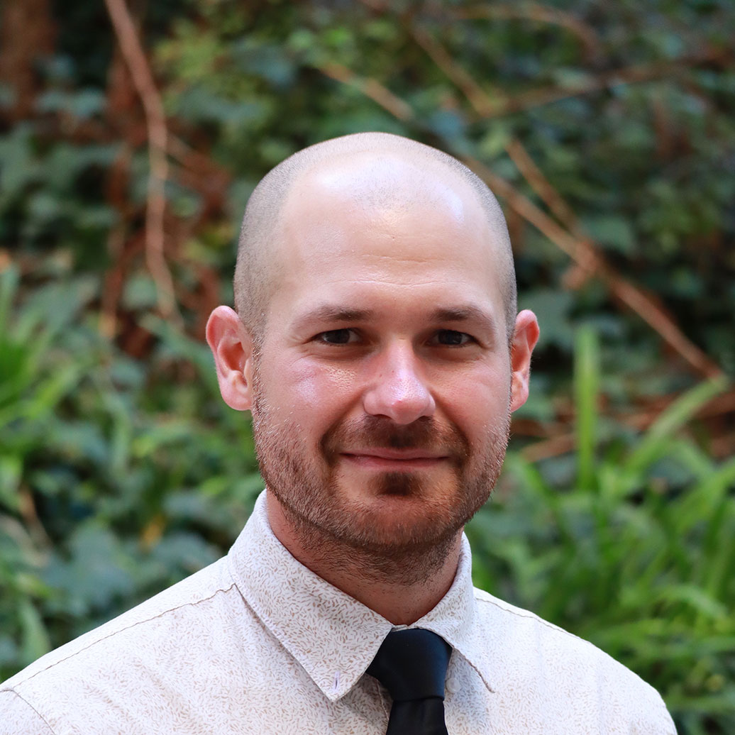 Headshot of scholar. Green plants behind scholar. Scholar is bald, has facial hair, and is wearing a white patterned button up shirt with a black tie