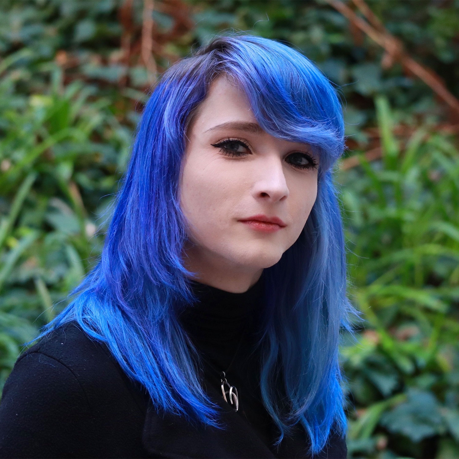 Headshot of scholar. Green plants behind scholar. Scholar has medium length bright blue hair, red lipstick. They are wearing a black long sleeve shirt and a necklace with a silver pendant. They have black eyeliner and red lips