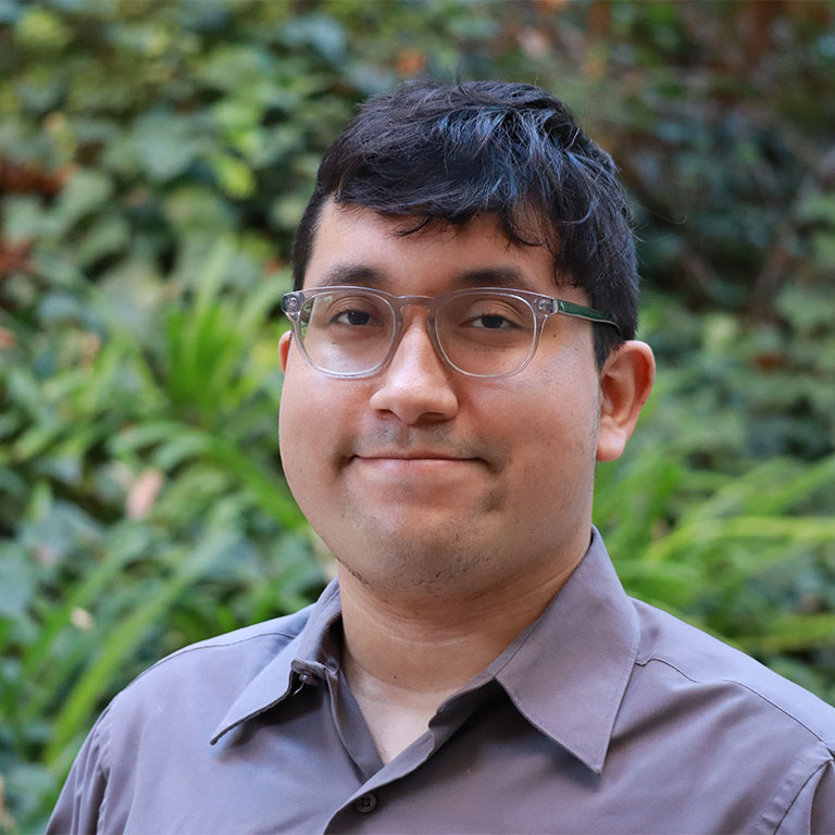 Headshot of scholar. Green plants behind scholar. Scholar has short black hair and is wearing clear reading glasses and a purple button up shirt