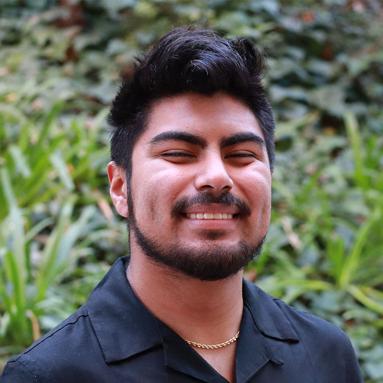 Headshot of scholar. Green plants behind scholar. Scholar has black hair and also facial hair. Wearing a black button up shirt with a gold chain necklace