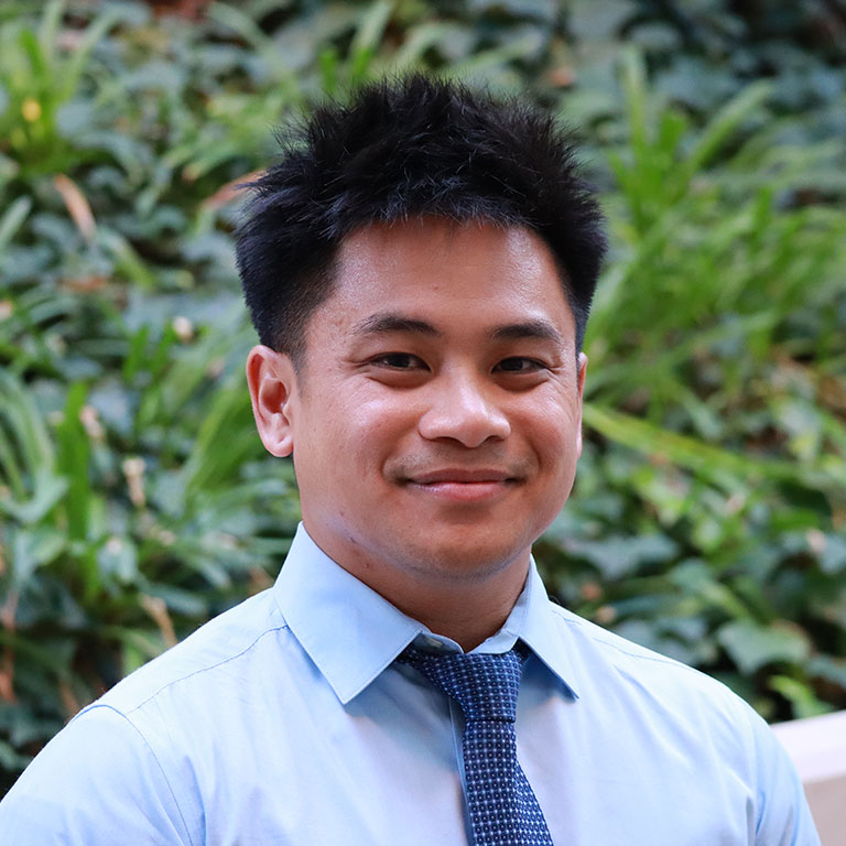 Headshot of scholar. Green plants behind scholar. Scholar has short black hair and is wearing a light blue button up shirt with a blue and white patterned tie