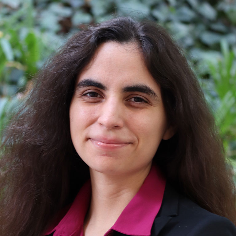 Headshot of scholar. Green plants behind scholar. Scholar has long black hair and wearing a bright pink button up shirt with a black blazer