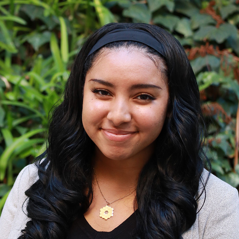 Headshot of scholar. Green plants behind scholar. Scholar has long black curly hair and wearing a black thick cloth headband. Is wearing a black shirt with a grey knitted blazer. Wearing a gold necklace with a gold pinwheel like pendant