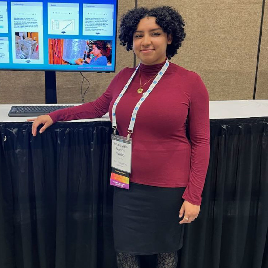 Scholar presenting at a Conference stands in front of computer. White name badge around the neck. Black curly hair. Red long sleeve shirt, black skirt, and black lace tights. Gold necklace
