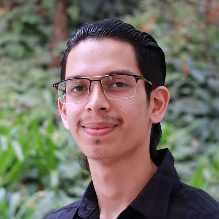 Headshot of scholar. Green plants behind scholar. Scholar has slicked back black hair and wearing a button up black shirt. Also wear square reading glasses with a black trim around glasses