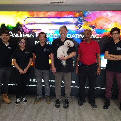Industry Assessment Center Team with 6 people featured. A colorful rainbow background is behind the people. From left to right there is one male, one female, and then 4 other males. The fourth male is holding a white dog. All are wearing black polos and one is wearing a red polo.