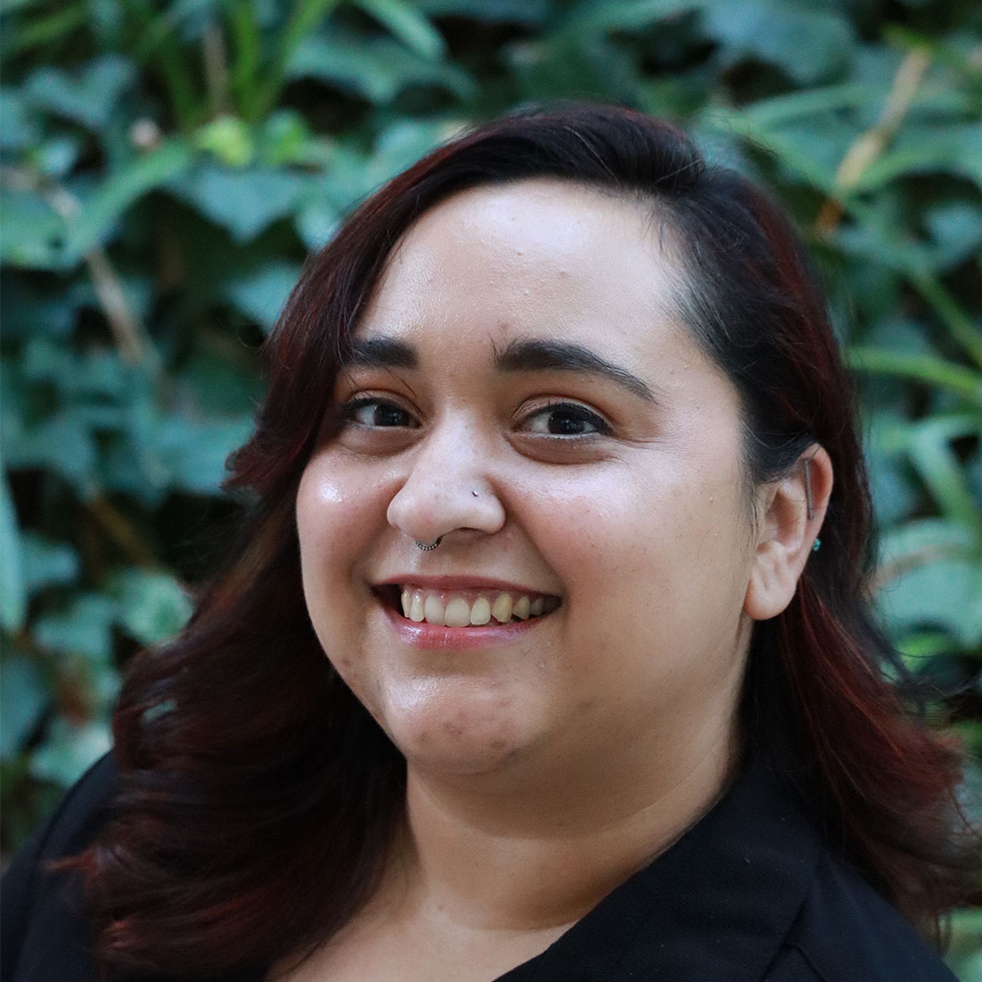 Headshot of scholar. Green plants behind scholar. Scholar has medium length dyed auburn hair. Wearing a button up black shirt. Has a loop nose earring and the top of her ear pierced with a bar and ball earring