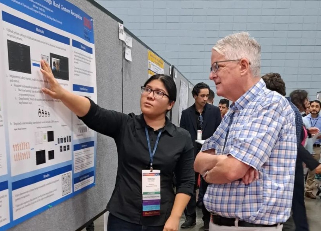 Scholar presents a poster for SACNAS conference in Puerto Rico. Wearing a conference name badge around her neck and a button up black dress shirt and glasses. Dean Roberts stands next to her