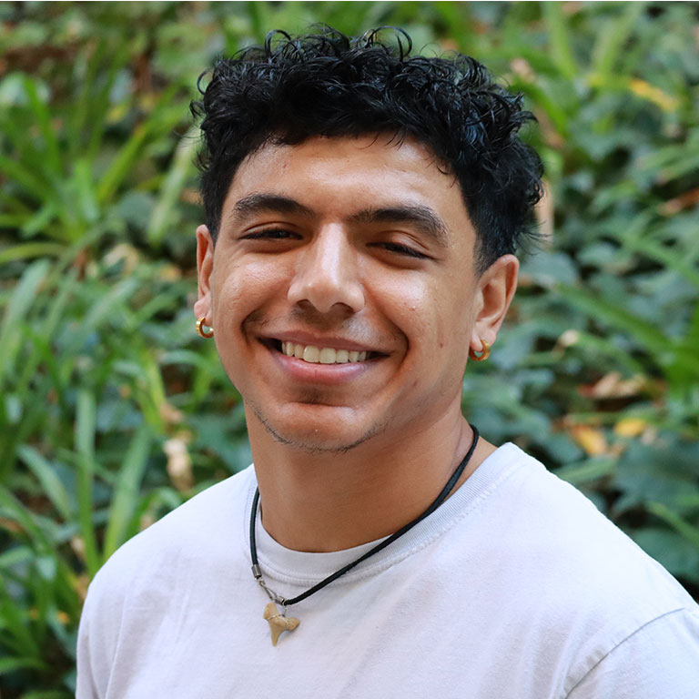 Headshot of scholar. Green plants behind scholar. Scholar has short curly black hair and wearing a white cotton t- shirt. Wearing a black necklace with a shark tooth pendant and two small gold earrings