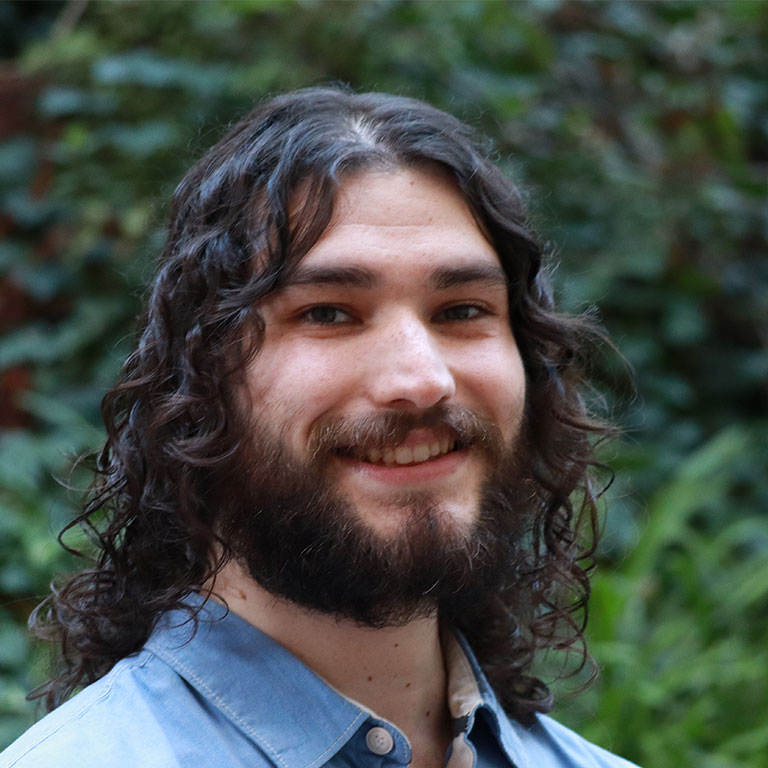 Headshot of scholar. Green plants behind scholar. Scholar has long curly black hair and facial hair. Wearing a light blue button up shirt