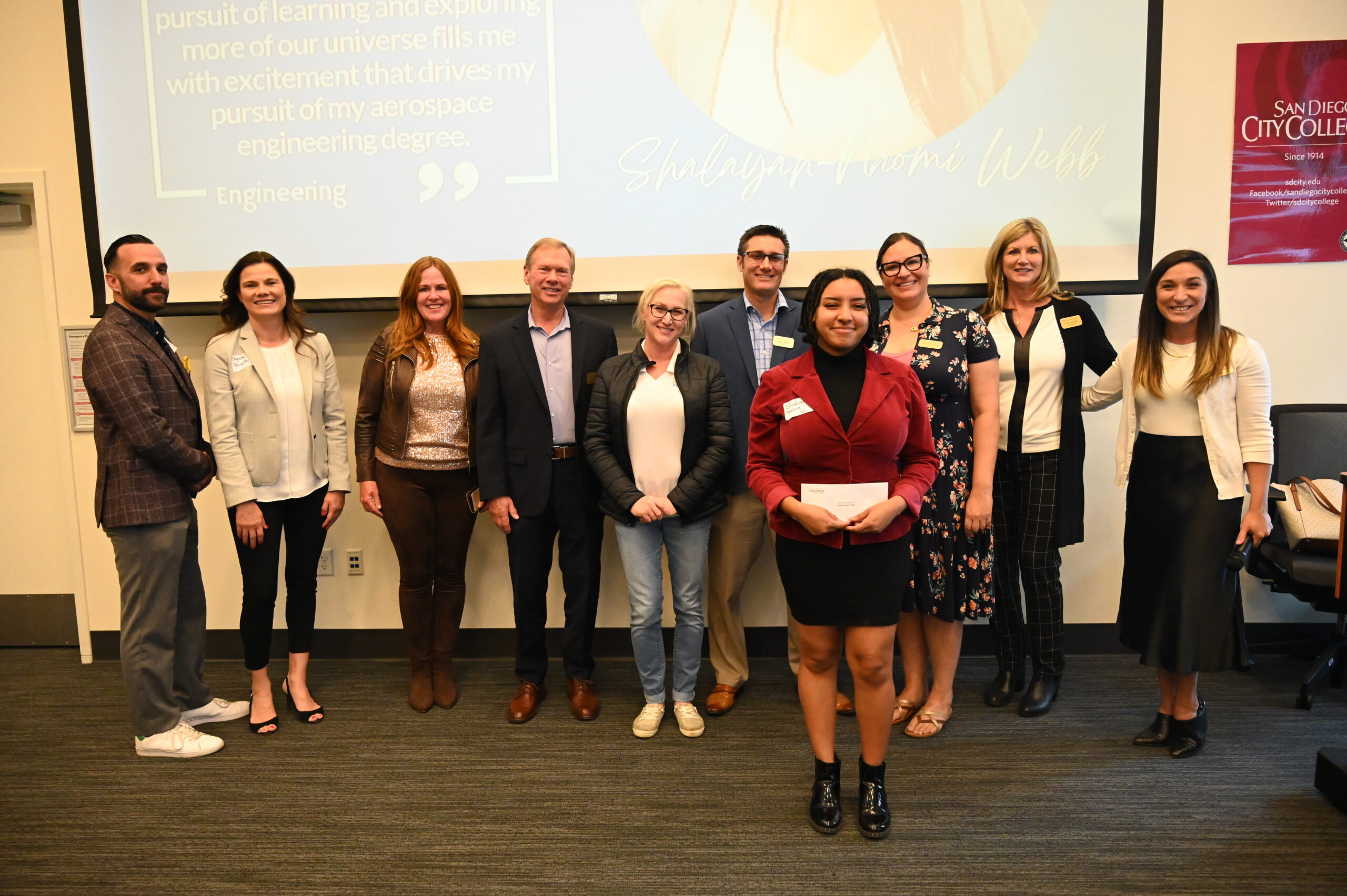 Scholar Naomi Webb stands in front of the Scholarship committee. There are 9 people behind Naomi. She has short braided black hair, a black shirt and skirt with black boots nd a red dress coat. She holds an envelope from the scholarship committee.