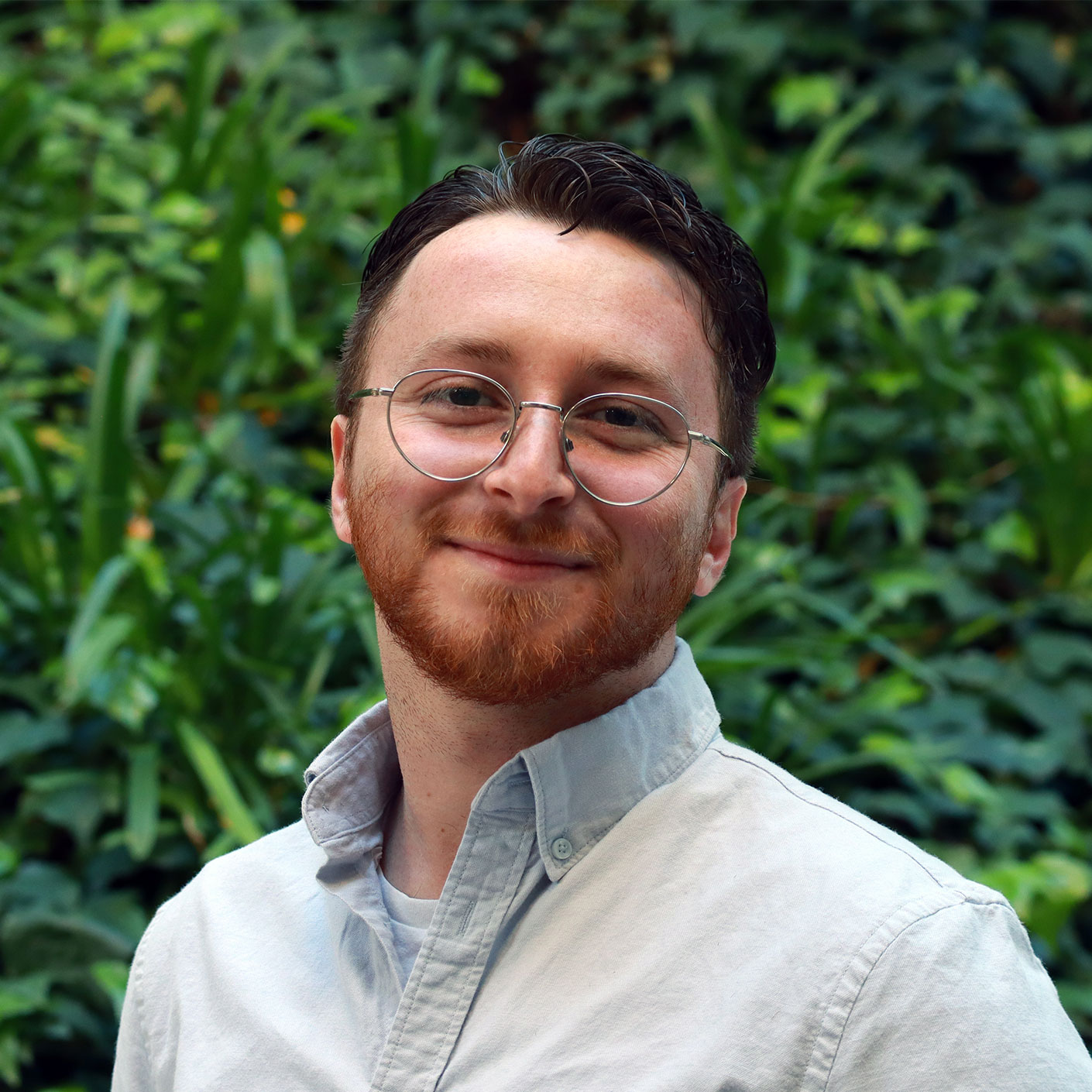 Headshot of scholar. Green plants behind scholar. Scholar has short brownish black hair. Wearing a light blue jean button up shirt. Underneath the button up shirt is wearing a white cotton shirt. Has clear round glasses and redish-brown facial hair that is cut into a clean beard