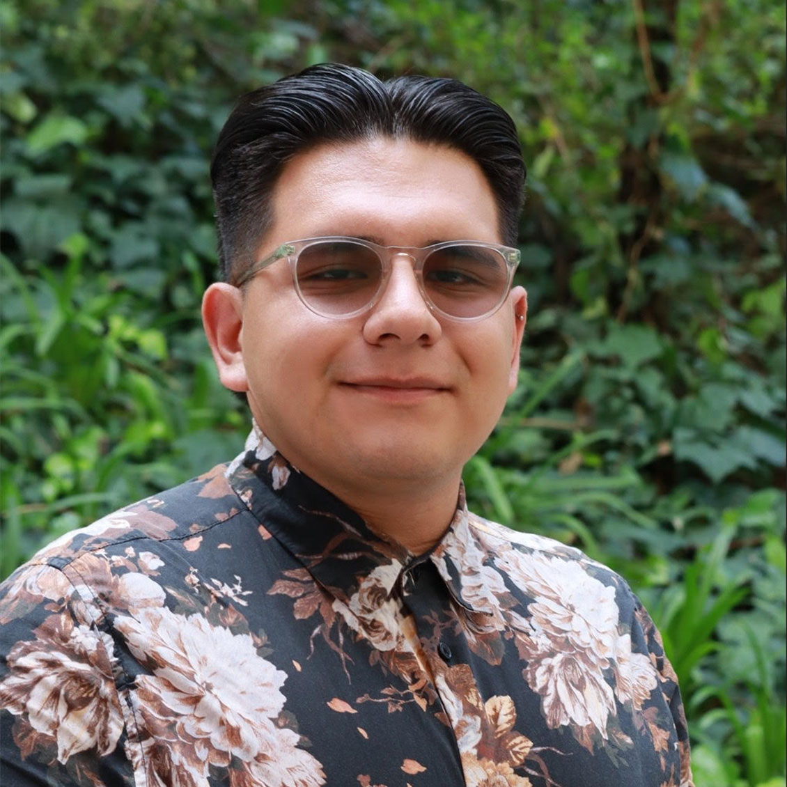 Headshot of scholar. Green plants behind scholar. Scholar has thick black short hair. Wearing a button up black floral shirt with brown and light brown flowers. Is wearing white shaded glasses that are oval shaped.