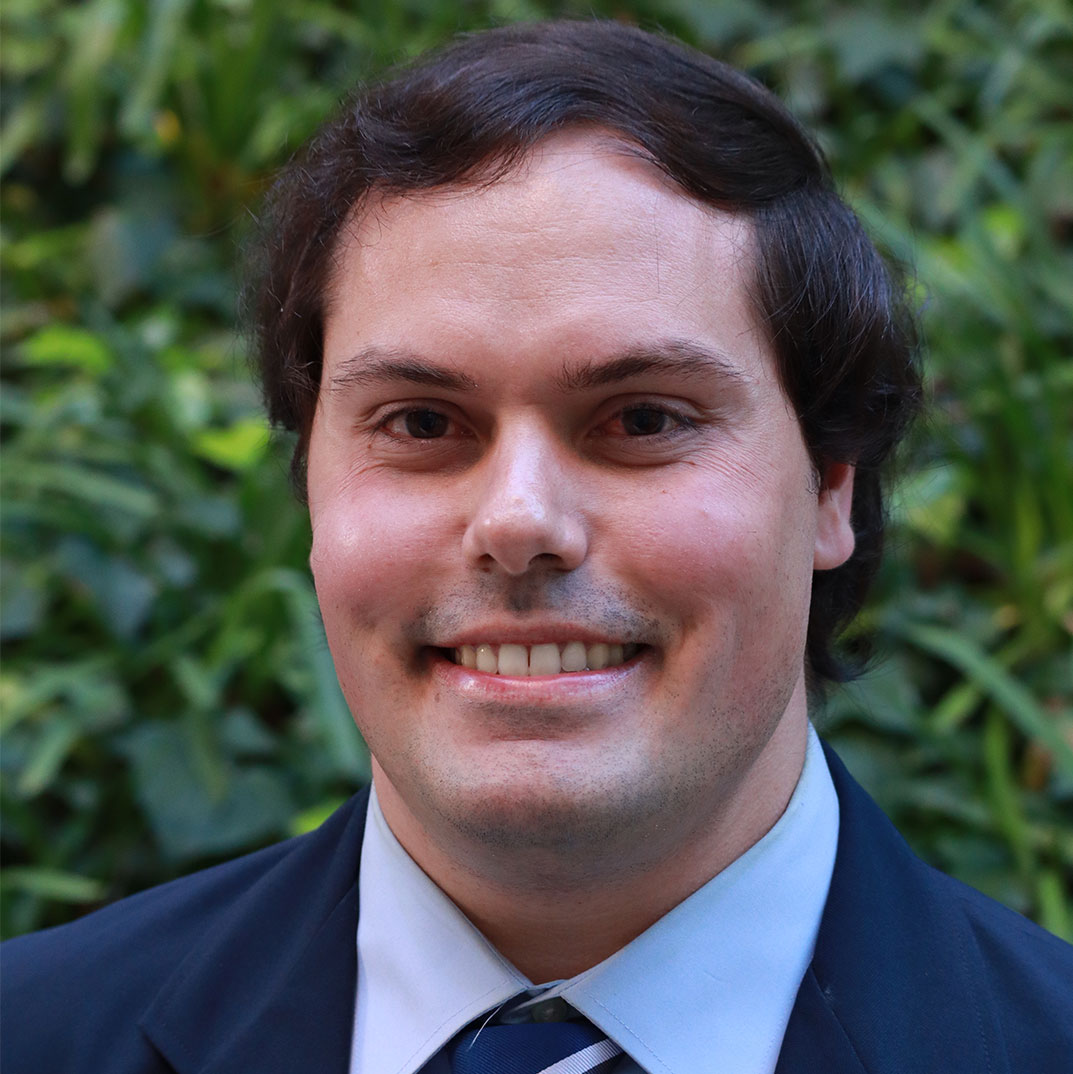 Headshot of scholar. Green plants behind scholar. Scholar has medium length brownish black hair. Top of hair curls over forehead. Wearing a dark blue suit jacket with a light blue button up shirt underneath and a striped tie with dark blue and light blue. Has a small light mustache.