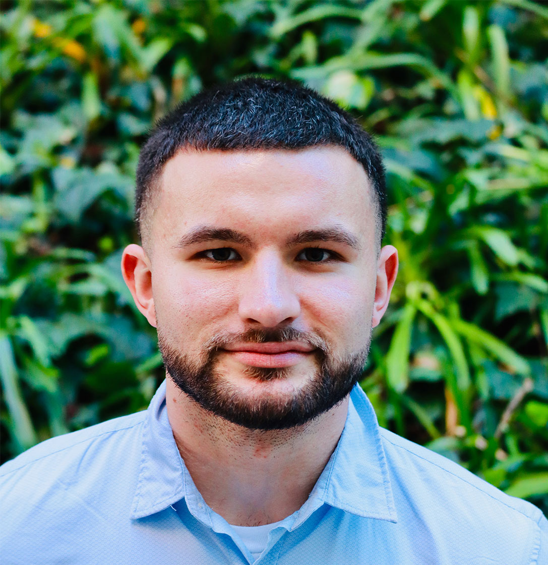 Headshot of scholar. Green plants behind scholar. Scholar has short black hair with some grey hairs. Has a black goatee and clean cut facial hair. Has some neck hair stubs. Is wearing a light blue button up shirt with a white cotton t-shirt underneath. They have thick black eyebrows.