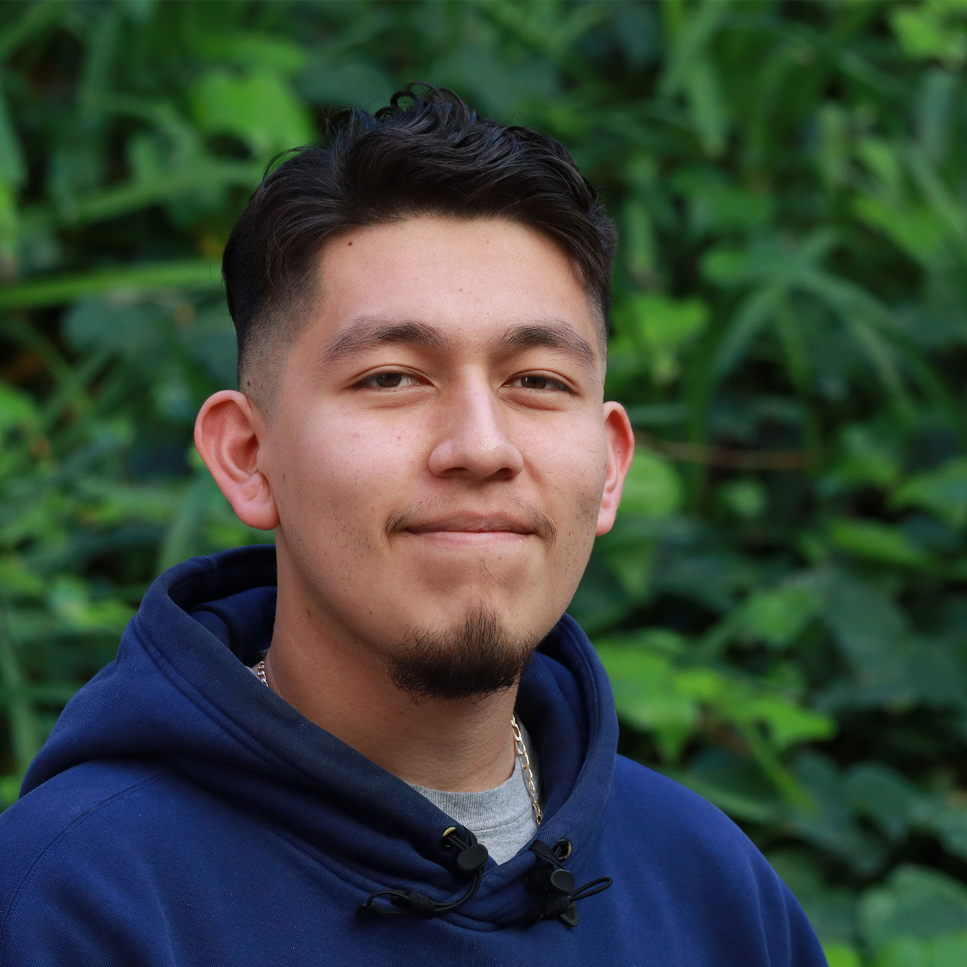 Headshot of scholar. Green plants behind scholar. Scholar has short black hair. Has a small black goatee. Wearing a blue sweater with a hood towards his back. Grey shirt inside hoodie. Silver chain around their neck.