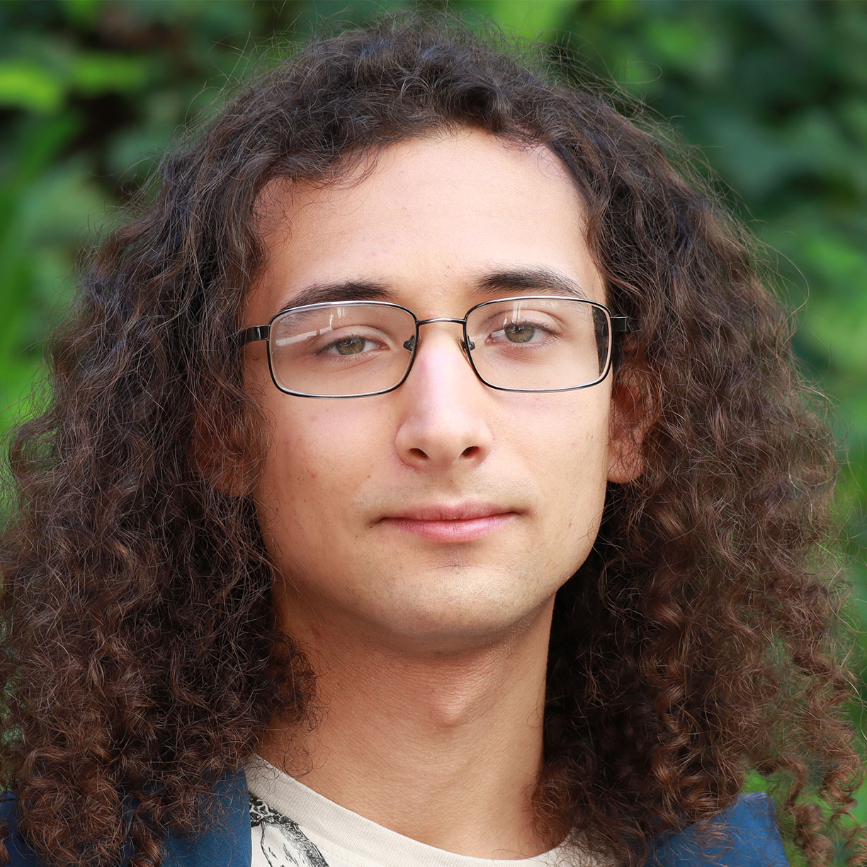 Headshot of scholar. Green plants behind scholar. Scholar has medium length curly brown hair. Wearing square clear glasses. Has hazel eyes and is smiling with no teeth a slight smile. Wearing a blue coat and light brown and black t- shirt inside jacket