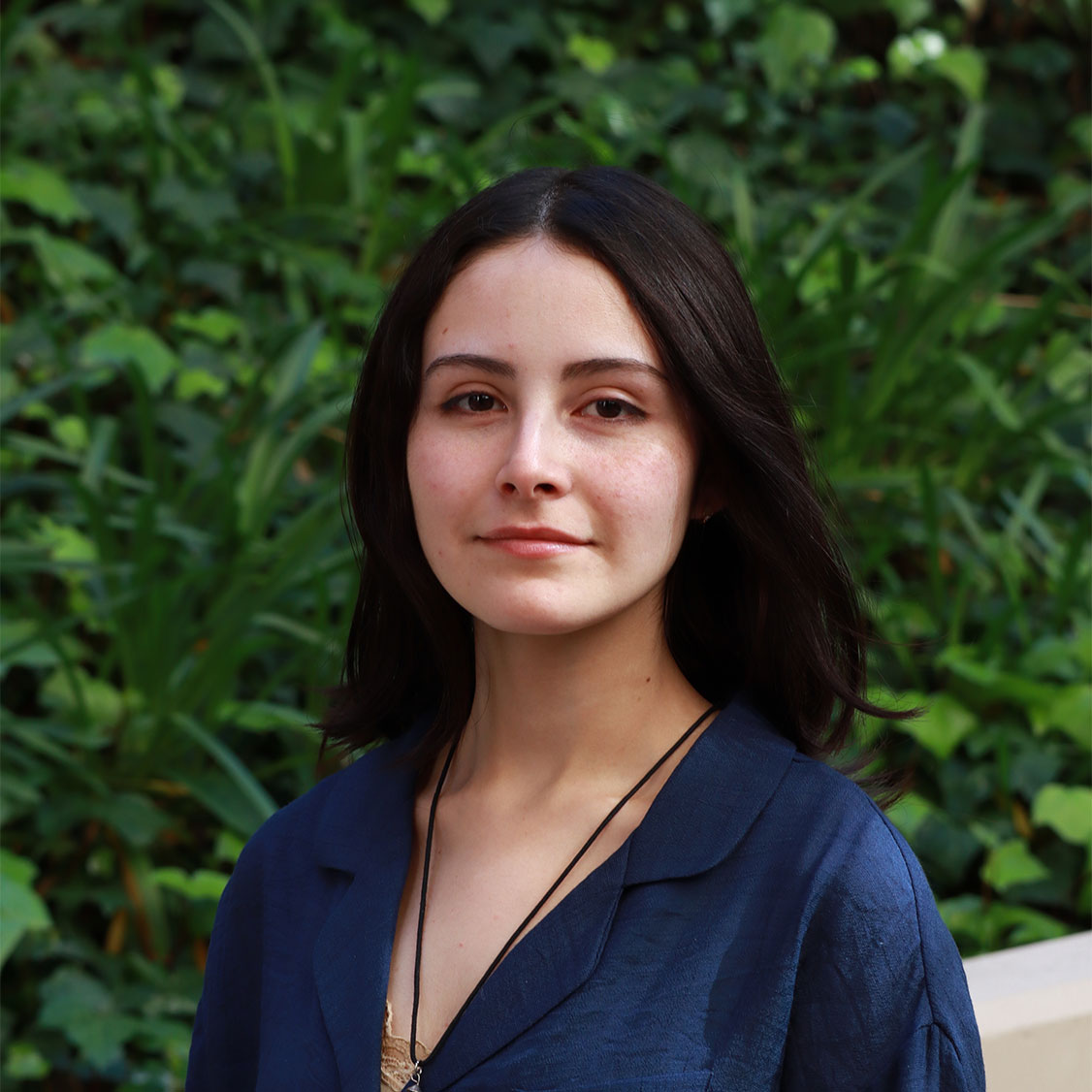 Headshot of scholar. Green plants behind scholar. Scholar has short straight hair to their shoulders. Clean shaped eyebrows with light soft pink eye shadow around the eyes and is wearing soft light pink lipstick. Wearing a dark blue dress shirt with long sleeves. Has gold lace camisole underneath the dark blue shirt. Has a long black string for a necklace.