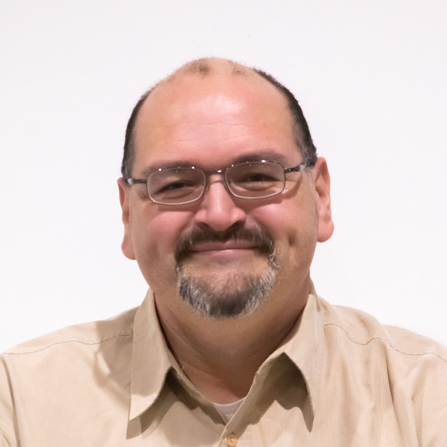 Headshot of scholar. Scholar is bald with small patches of black hair on top and on side. Has a grey and white goatee and 1 dimple. Smiling with no teeth. Wearing a brown button up shirt with a white cotton undershirt underneath