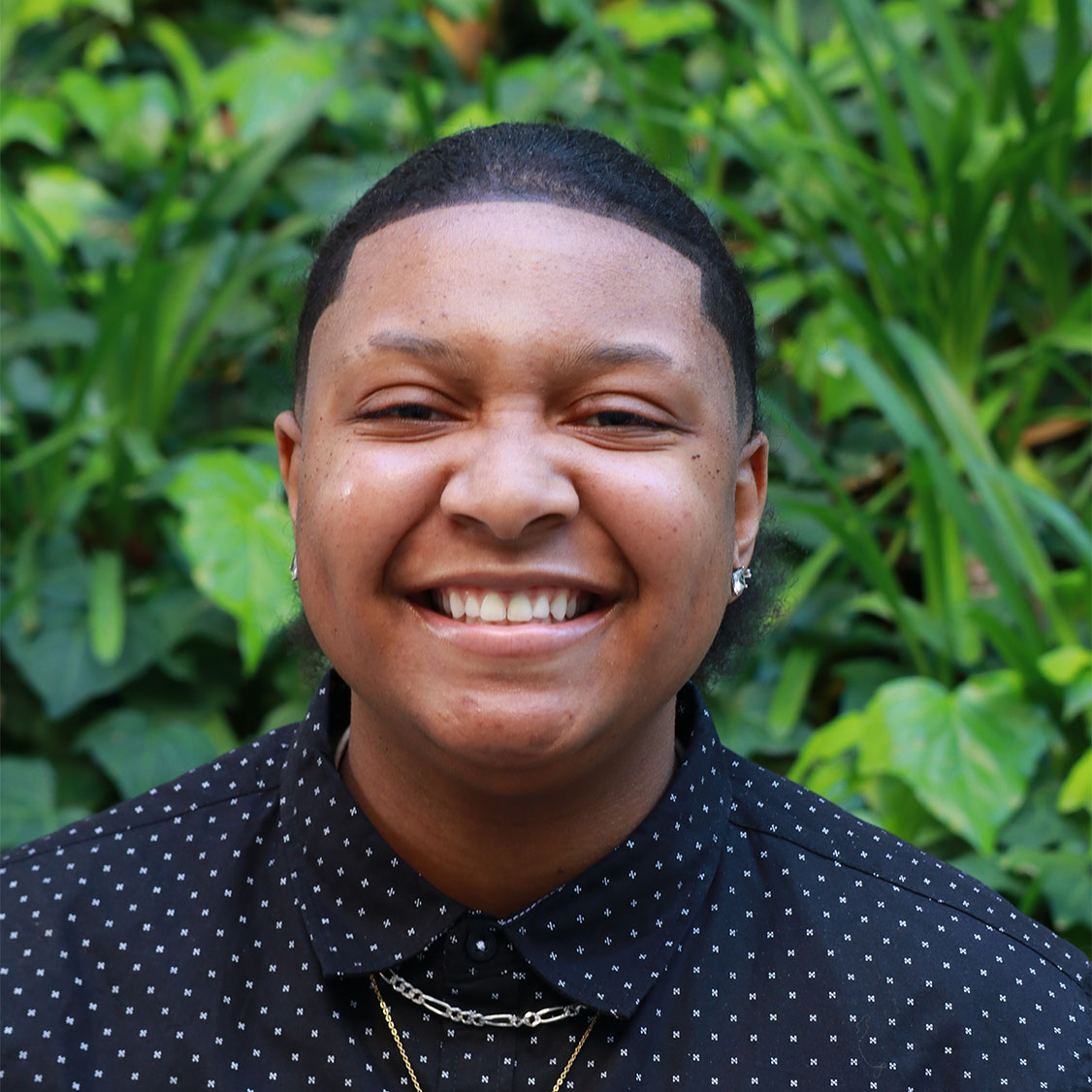 Headshot of scholar. Green plants behind scholar. Scholar has short black clean cut buzzed hair. Has a big smile with teeth. Has thin black eyebrows. Wearing a diamond stud earring on one ear. Wearing a button up dark blue with white title patterns throughout shirt. Has a silver thicker chain necklace and a thin longer gold chain