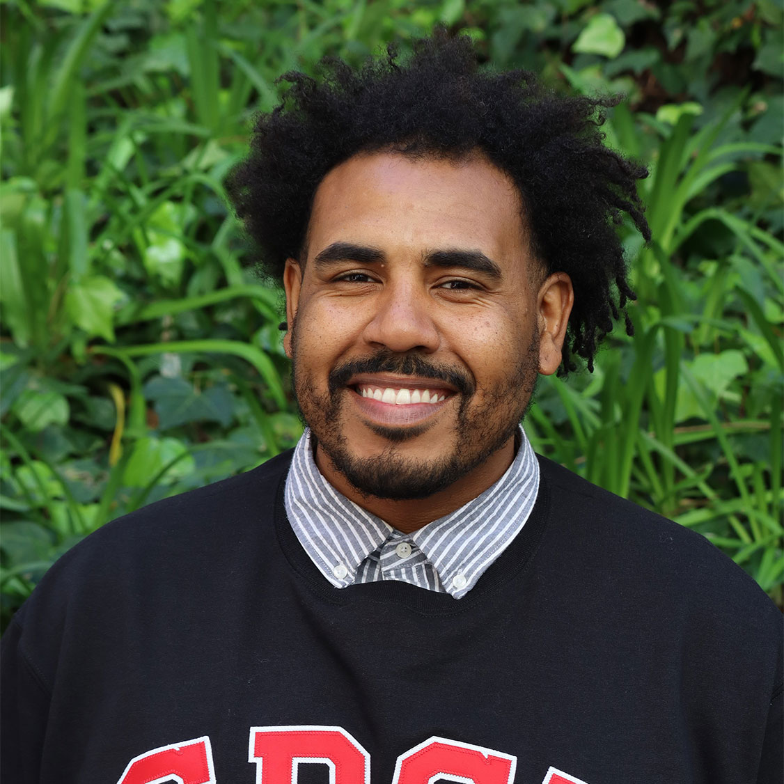 Headshot of scholar. Green plants behind scholar. Scholar has black medium size dreads for hair. Scholar is tall has thick black eyebrows and black facial hair on his cheeks. The facial hair connects as a goatee on the face. Wearing a striped blue and white button up inner shirt. On top of the inner shirt they wear a black SDSU sweater. SDSU is in red letters on the sweater