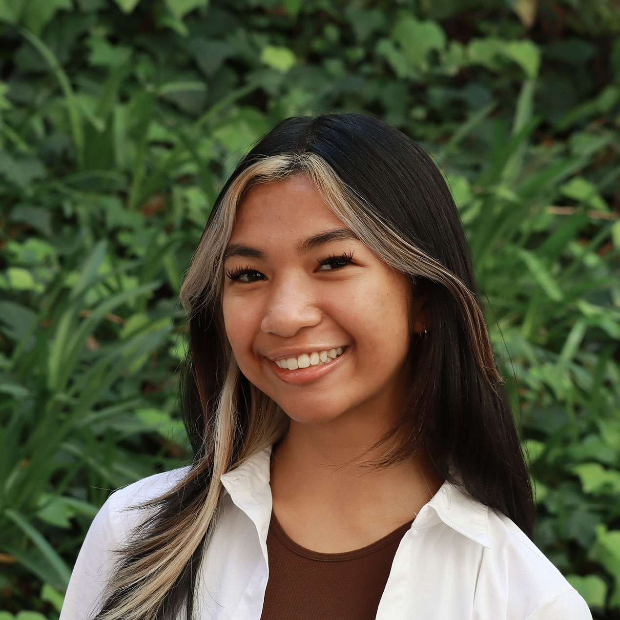 Headshot of scholar. Green plants behind scholar. Scholar has medium black length layered hair with bangs split to the side. The bangs are blond highlights. Is smiling with teeth and is wearing soft brown lipstick with lipgloss. Wearing a white button up shirt and a brown shirt is underneath the white shirt. Has long black lashes.