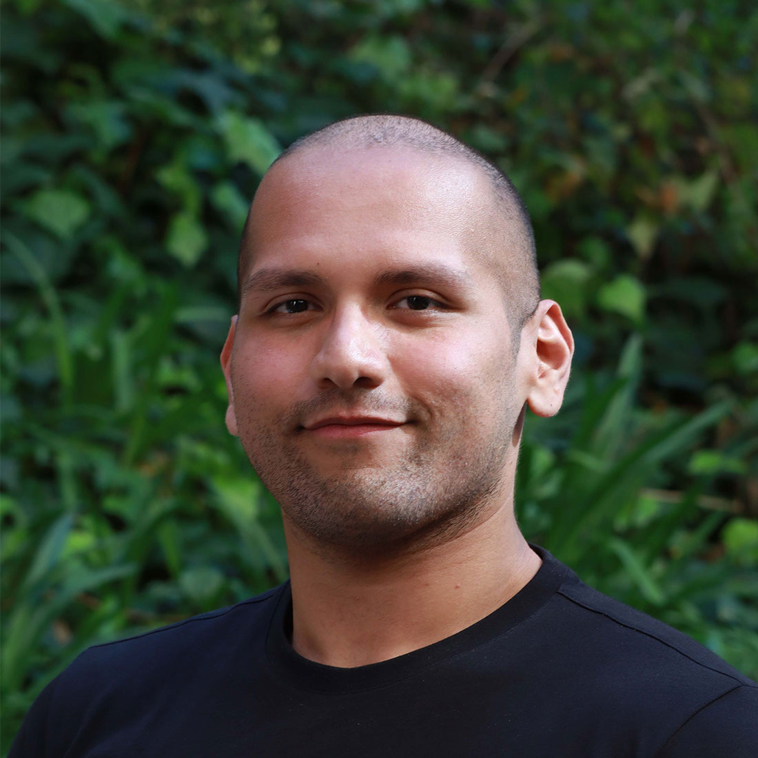 Headshot of scholar. Green plants behind scholar. Scholar has a bald head with short black hair that is buzzed. They have thick black eyebrows. Black stubby facial hair on face. Wearing a black shirt.