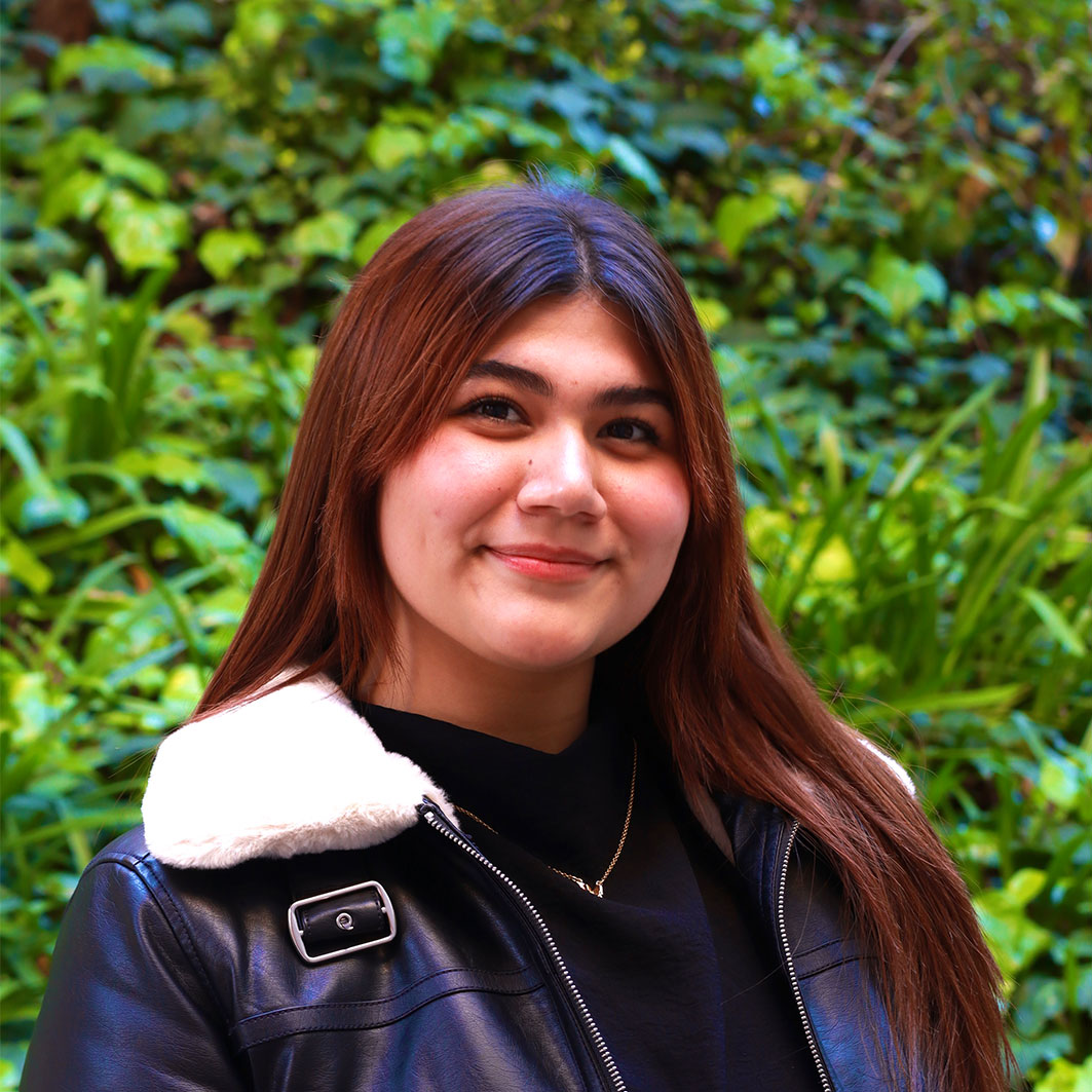 Headshot of scholar. Green plants behind scholar. Scholar has medium brownish red length layered hair with bangs split to the side. Is smiling with no teething wears soft red lipstick. Wearing a black leather jacket with a white fur color. Wears a black shirt inside and has a gold necklace around their neck