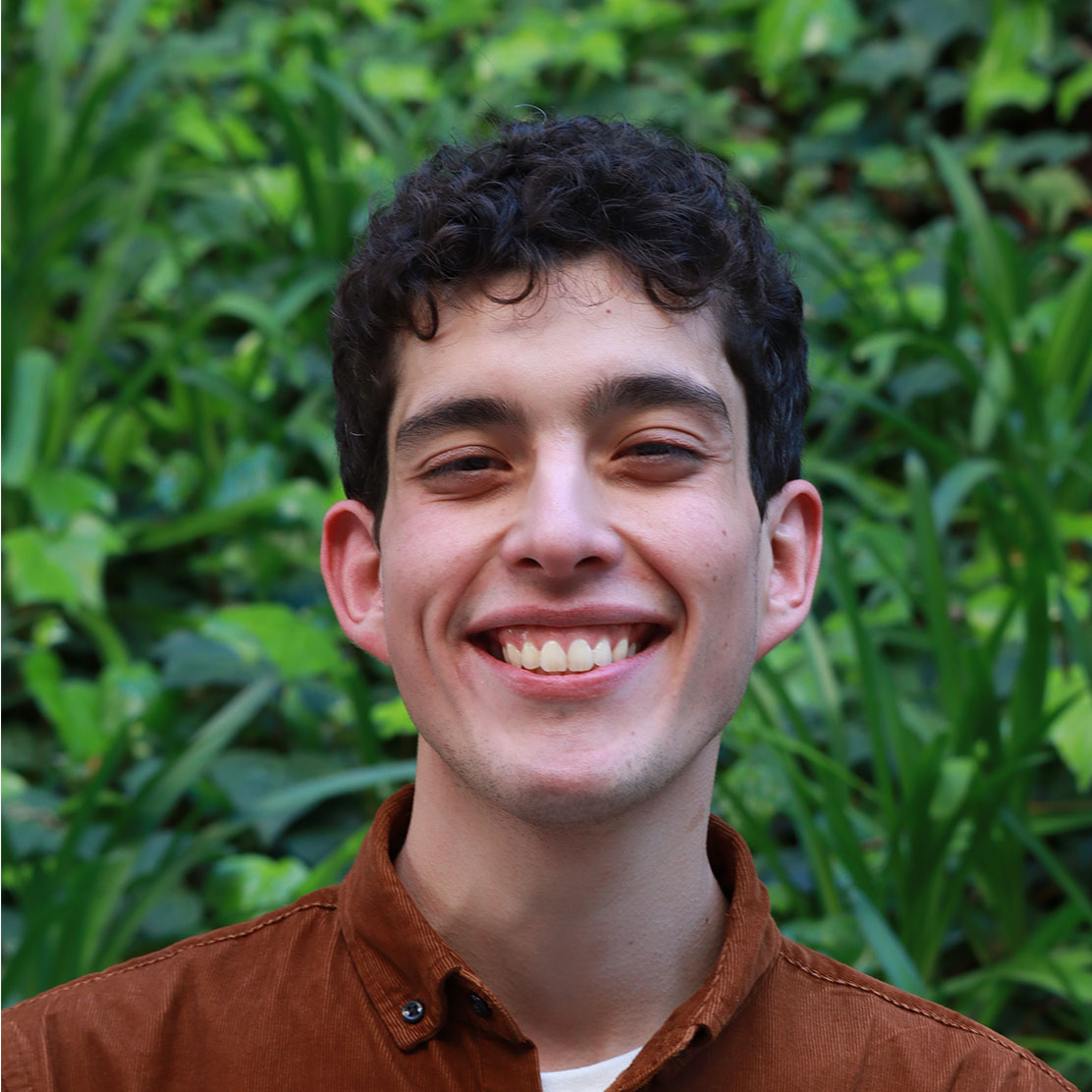 Headshot of scholar. Green plants behind scholar. Scholar has short clean cut black hair with curly bangs. Has thick black clean eyebrows. The Scholar is smiling big with teeth. They are wearing a brown button up shirt with a white cotton shirt underneath the brown shirt