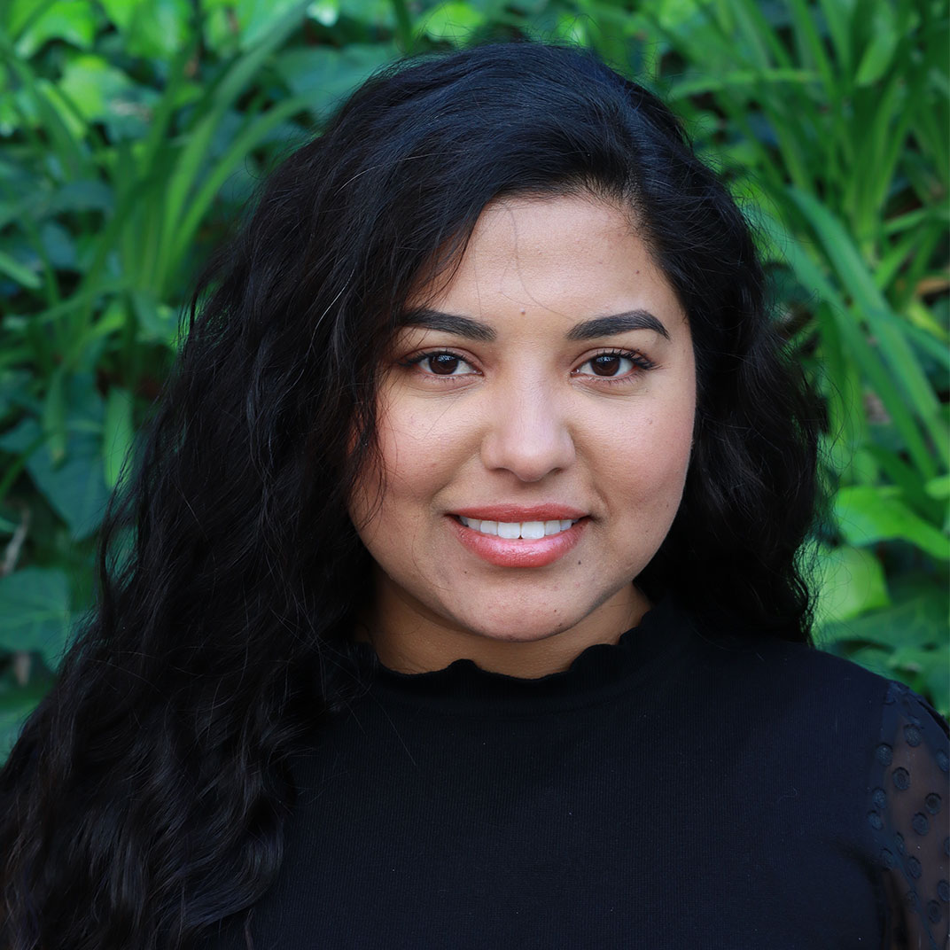 Headshot of scholar. Green plants behind scholar. Scholar has long think wavy black hair that is hanging over their shoulder. The scholar has nicely shaped eyebrows and is wearing a black ruffled shirt. Has a big smile with teeth showing and is wearing light pink lipstick.