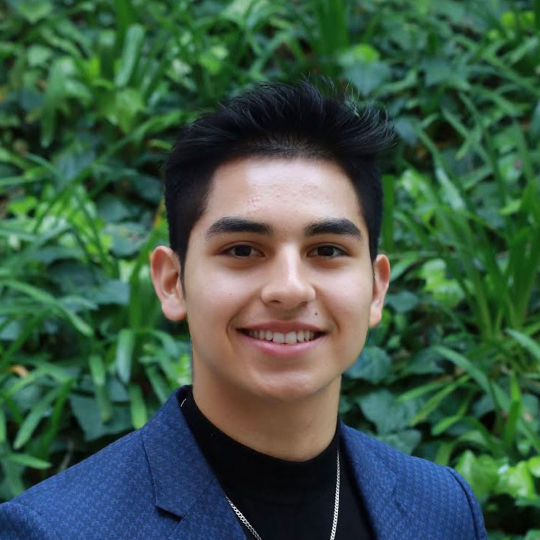 Headshot of scholar. Green plants behind scholar. Scholar short black hair with bangs that go up. Thick eyebrows. Is smiling with teeth. Has a tiny mole above their lip. Wearing a dark blue patterned suit with a black undershirt underneath. Wears a silver chained necklace.