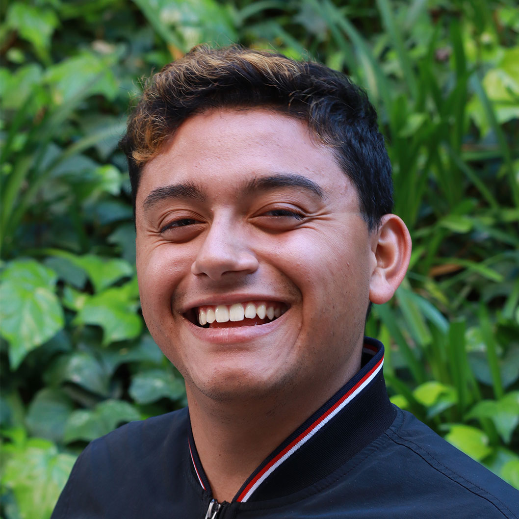 Headshot of scholar. Green plants behind scholar. Scholar has short black hair with brown highlights. Has a big smile and is smiling with teeth. Is wearing a navy jacket with a white and stripe around the collar.