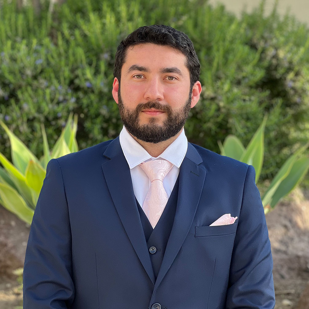 Headshot of scholar. Green plants behind scholar. Scholar has short black wavy hair. Has thick black eyebrows. The Scholar is smiling no teeth. They are wearing a dark blue two piece suit with a white button up shirt underneath the suit. Is wearing a light pink tie and a light pink pocket square coming out of the pocket.