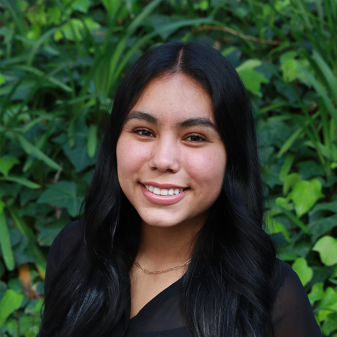 Headshot of scholar. Green plants behind scholar. Scholar has long black curled hair parted in the middle of face. They are wearing a black silk button up shirt with a black shirt underneath. Has light pink lipstick and is smiling with teeth. Wears a gold chained necklace