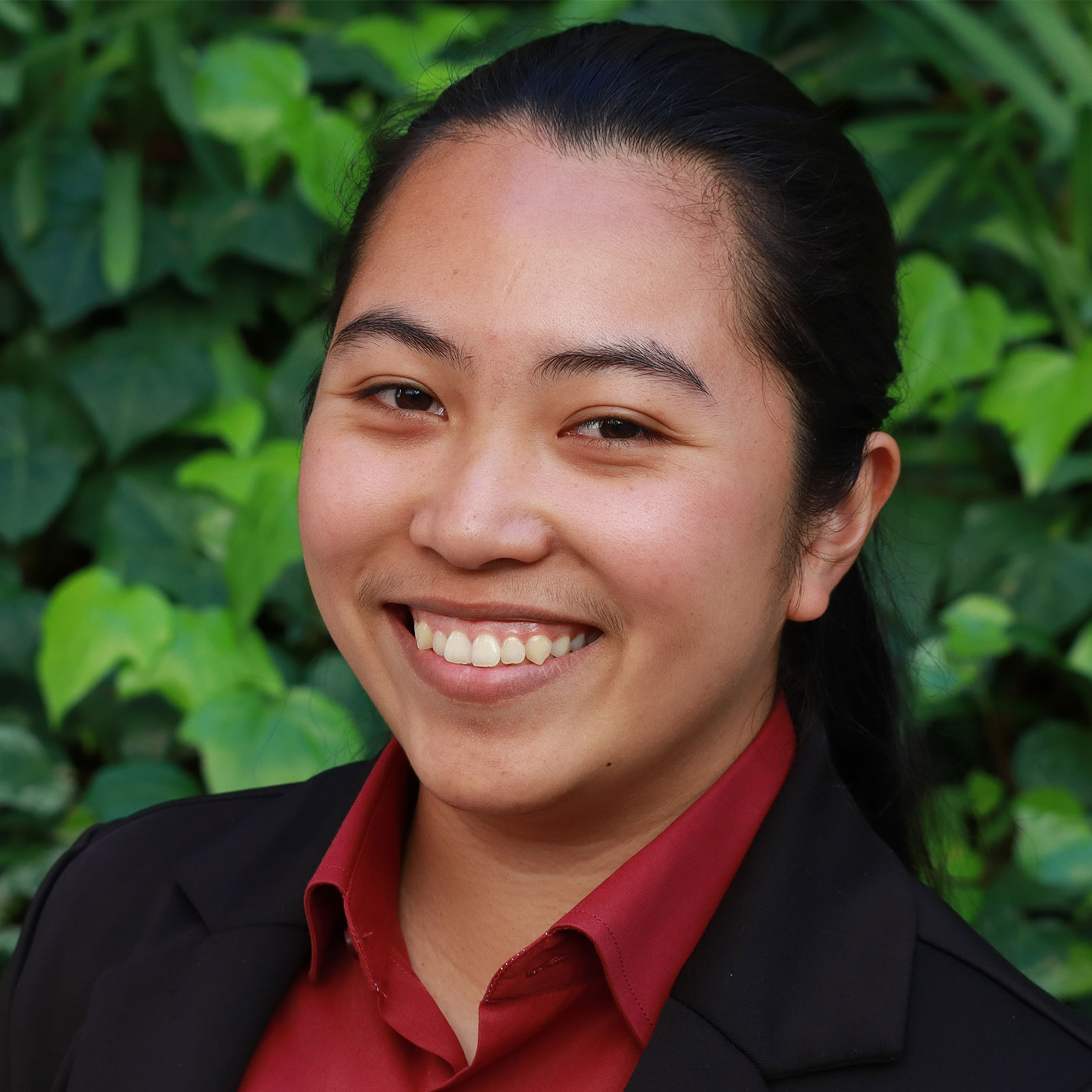 Headshot of scholar. Green plants behind scholar. Scholar has black hair that is pulled back in a pony tail. Has thick eyebrows. Wearing a black dress coat with a red button up shirt underneath. Is smiling big with teeth.