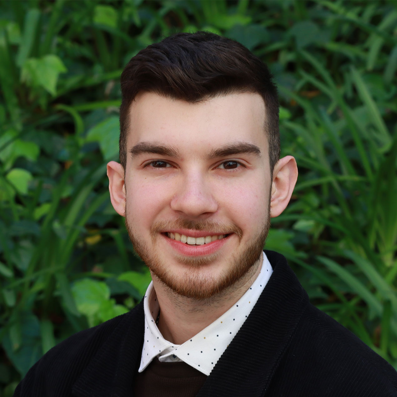 Headshot of scholar. Green plants behind scholar. Scholar has short thick wavy brown hair. Has thick brown eyebrows. The Scholar is smiling with teeth. Has a goatee and facial hair on their face. Has hair on their neck. Wearing a black sweater with a button up white shirt with black and brown polka dots