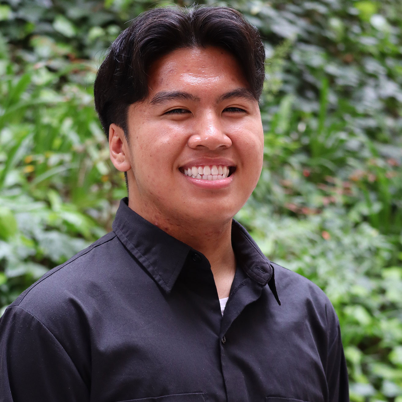 Headshot of scholar. Green plants behind scholar. Scholar has short black hair with thick bangs split in the middle. Wearing a black button down shirt. with a white undershirt. Has a big smile