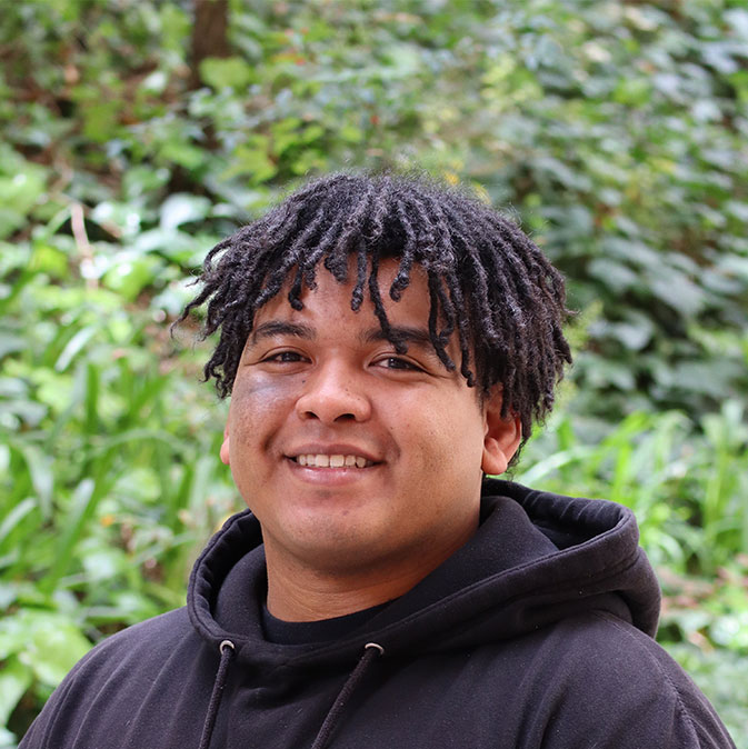 Headshot of scholar. Green plants behind scholar. Scholar has medium black hair that is dreaded. Hair length comes hangs down by eyes. Is wearing a black hooded sweater with the hood hanging on the back of their neck. Smiling with teeth. Has a color fragment on his right cheek.