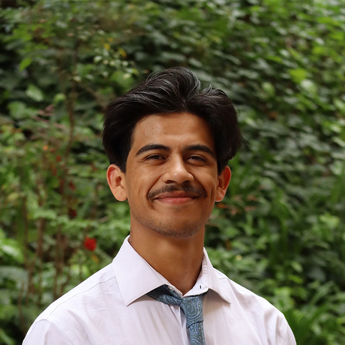Headshot of scholar. Green plants behind scholar. Scholar has short black wavy hair. Is wearing a white button up collar shirt with a blue patterned tie. Has a big smile with a mustache.