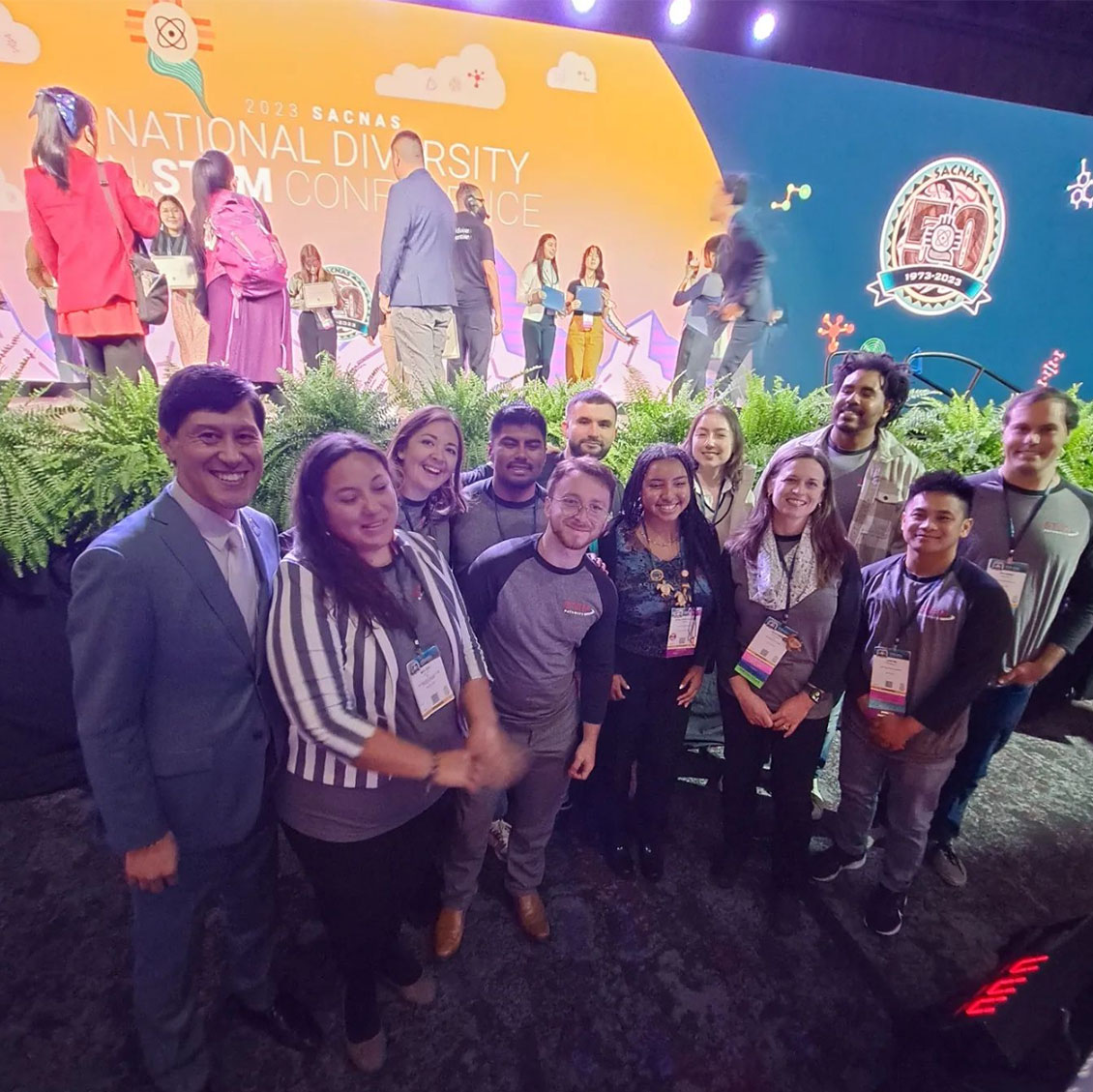 2023 N Di STEM SACNAS Conference. 12 people from the STEM Pathways Program both staff and scholars stand in front of main stage with their SACNAS conference badges hanging from their neck.The background is of diverse people coming together in front of the SACNAS logo 50th anniversary 1973- 2023.
