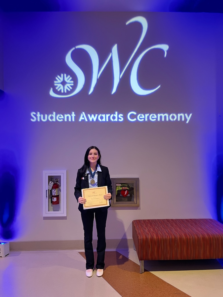 STEM Pathway Scholar Daniela Santibanez wears a black pantsuit with a light blue collar button up shirt, white dress shoes. Around her neck she wears a medallion and holds up her award for winning the Student Distinction Award (SODA) at Southwestern College in the spring of 2024. This award is considered the highest award granted to a Southwestern College Student. Only 20 students are selected each year! She stands in front of a SWC sign that says Students Awards Ceremony.