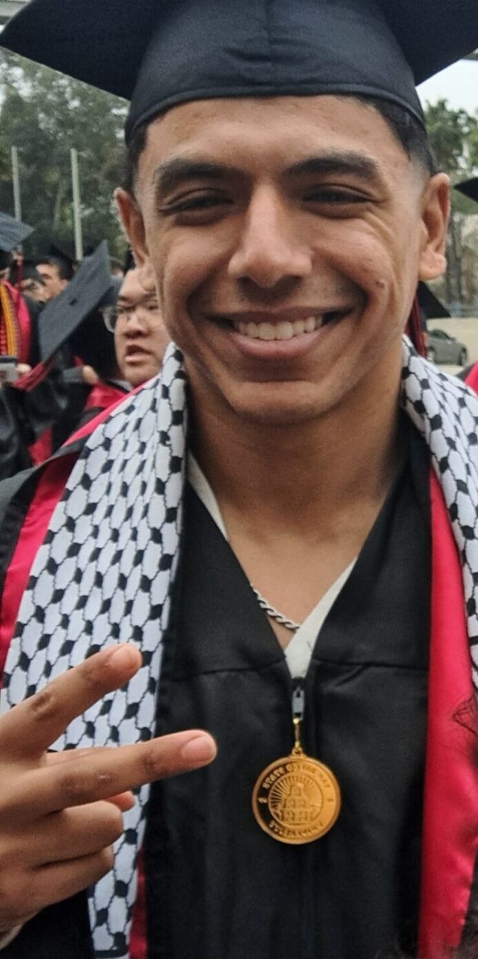 STEM Pathways Scholar dressed in graduation black cap, SDSU medalion, and graduation sashes smiles big holding up a peace sign graduates from SDSU in May of 2024.