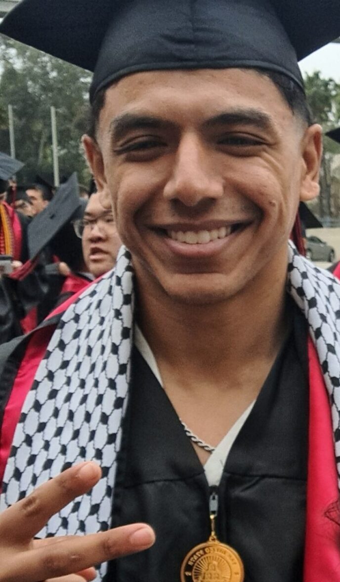 STEM Pathways Scholar Shaddy Elhamad is dressed in a graduation black cap, SDSU medalion, and graduation sashes smiles big holding up a peace sign as graduates from SDSU in May of 2024.
