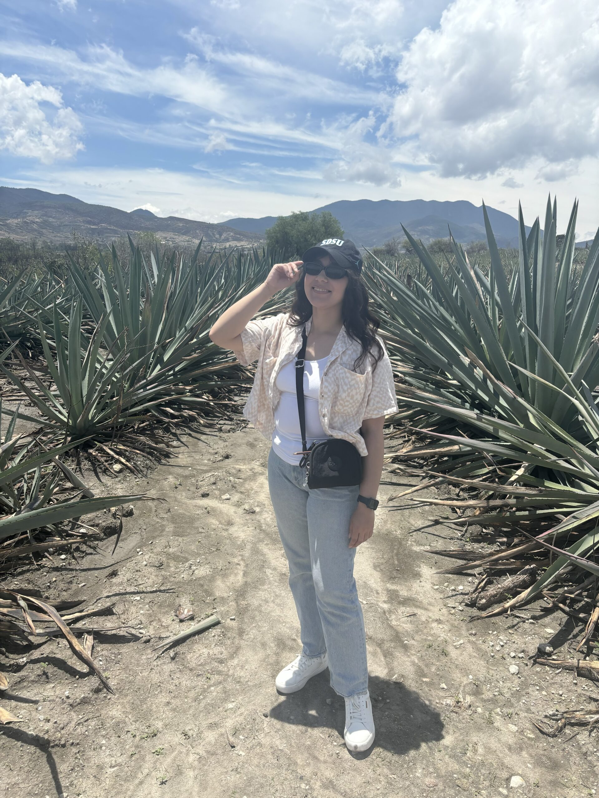 Scholar stands in between plants with a black and white SDSU cap and glasses. She tips her hat! Clear blue skies with the mountain is in the background. Scholar wears a white shirt with a patterned yellow shirt on top. She has white shoes and a balck watch and her black purse hangs over her shoulder. This picture was taken in Oaxaca, Mexico.