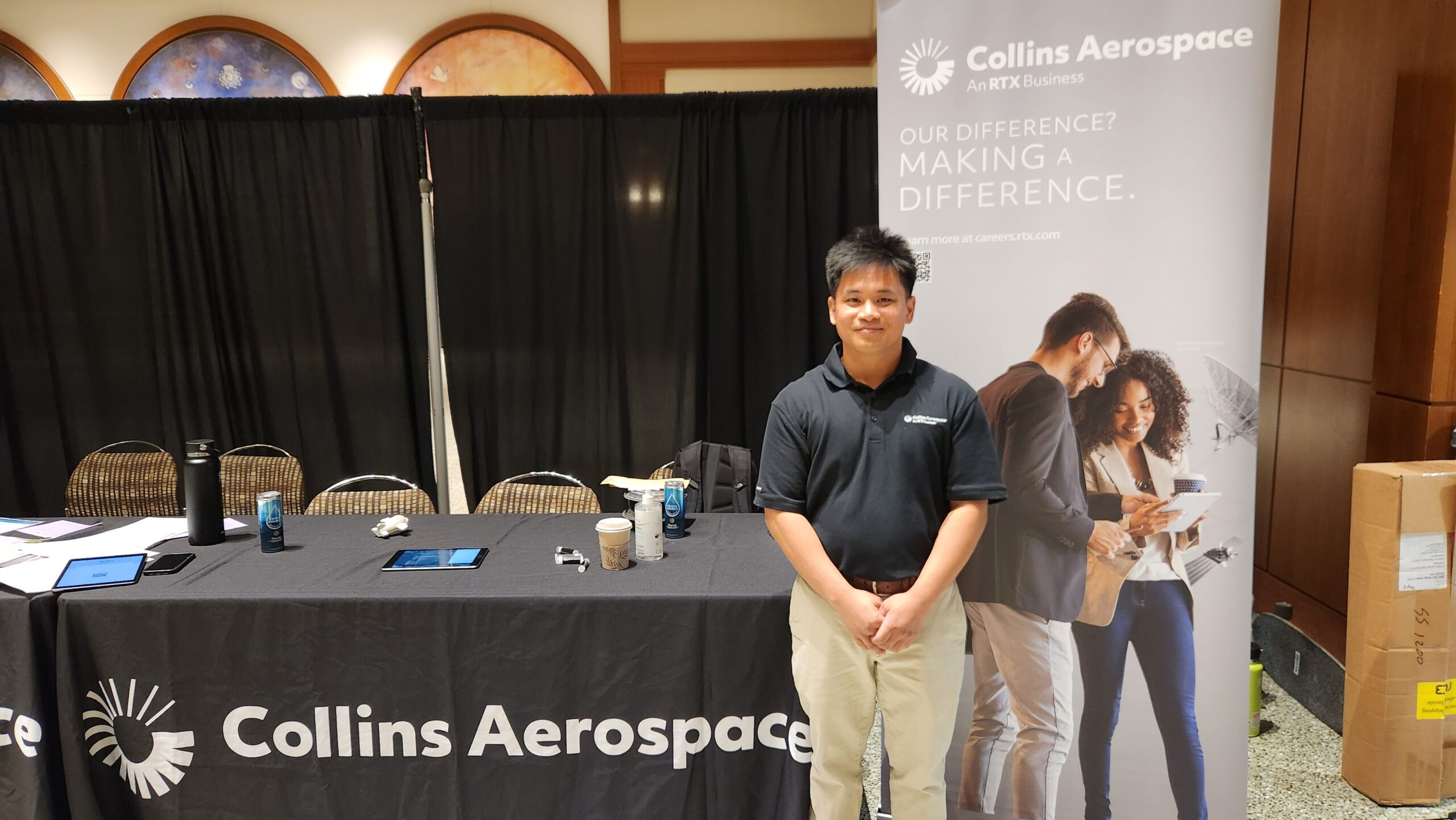 STEM Pathways Scholar is tabling at the Collins Aerospace table. Scholar stands in front of the table wearing a black polo from Collins Aerospace and brown khakis.