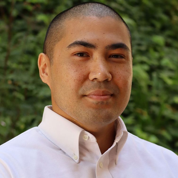 Aaron-newpsd-copy Headshot of scholar. Green plants behind scholar. Scholar has short buzzed black hair. They are wearing a white button up shirt.